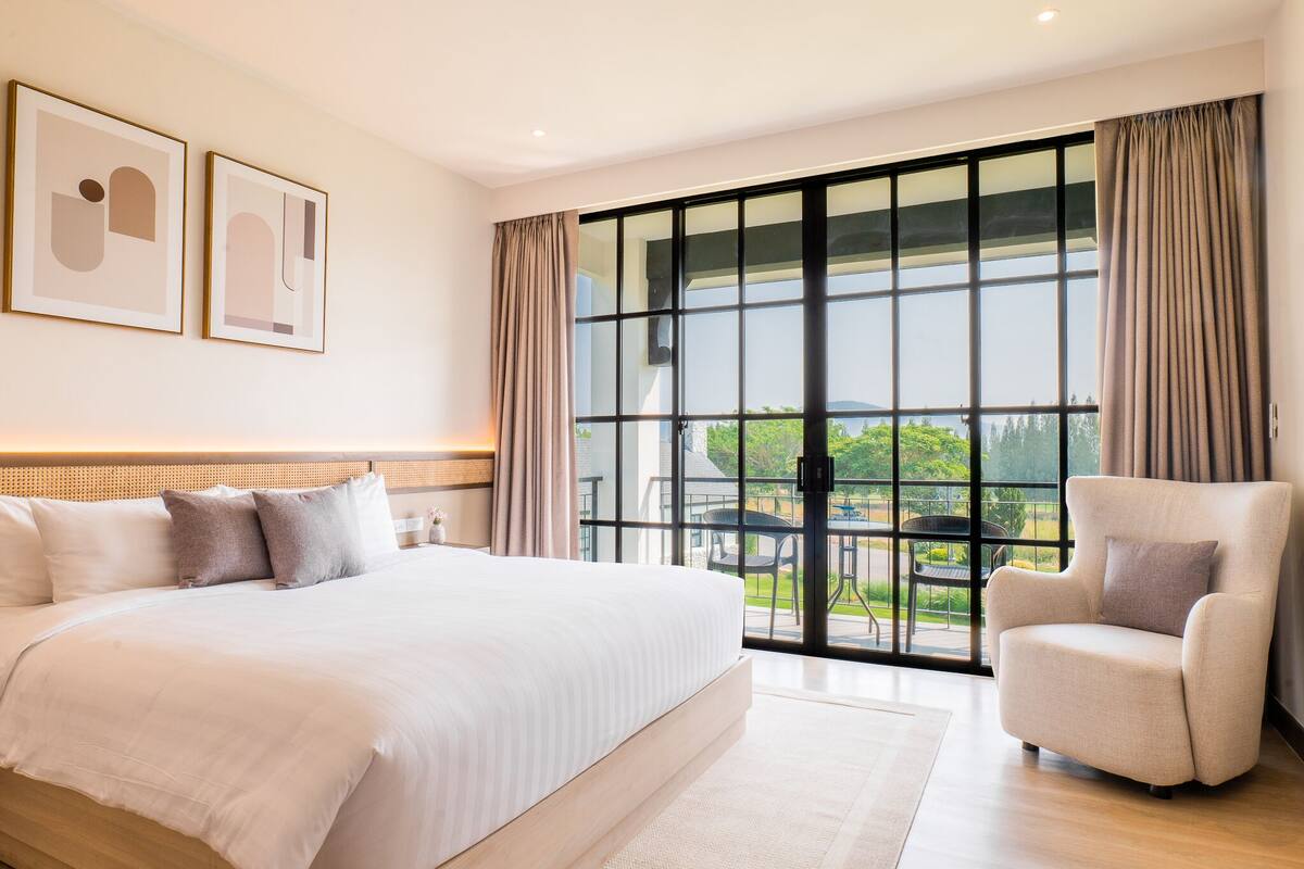 A modern bedroom with a large bed, a chair, and a view of the outdoors through a sliding glass door.