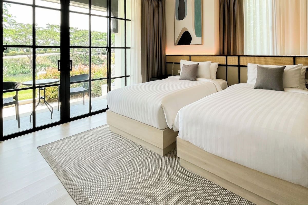 A hotel room with two beds, a large sliding glass door to a balcony, and a view of greenery outside.