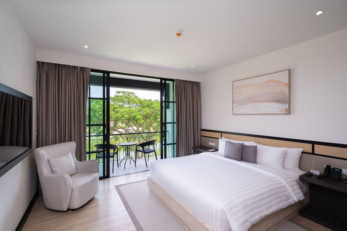 A modern hotel room with a large bed, a sofa, a small table, and a balcony with a view of greenery.