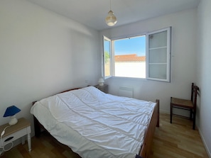A bedroom with a bed, a chair, a desk, and a window with an open shutter.