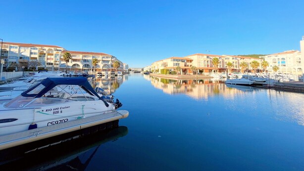 A marina with boats docked, a clear blue sky, and buildings with balconies and palm trees.