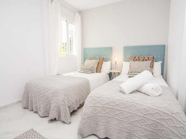 Habitación de hotel con dos camas, cada una con una colcha y almohadas estampadas, una mesita de noche pequeña con una lámpara y un ventanal con cortinas.