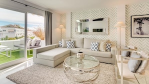 A modern living room with a glass coffee table, a sofa with patterned cushions, a large mirror, and a floor lamp. - Beautiful 3-bedroom apartment with terrace in Nueva Andalucía, Marbella (Marbella)