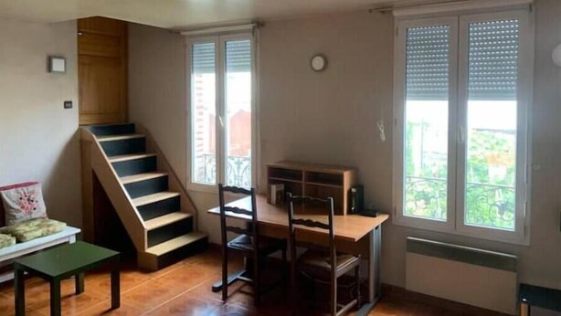 Een kamer met een houten trap, een eettafel met stoelen en grote ramen met jaloezieën.