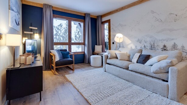 Un salon chaleureux avec un canapé, un fauteuil, une table basse et un téléviseur. On y trouve des rideaux, une lampe et un mur orné d’une fresque représentant une montagne.