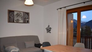 Ein Zimmer mit grauem Sofa, einem Holztisch und Stühlen. Ein Fenster bietet Blick auf Berge.