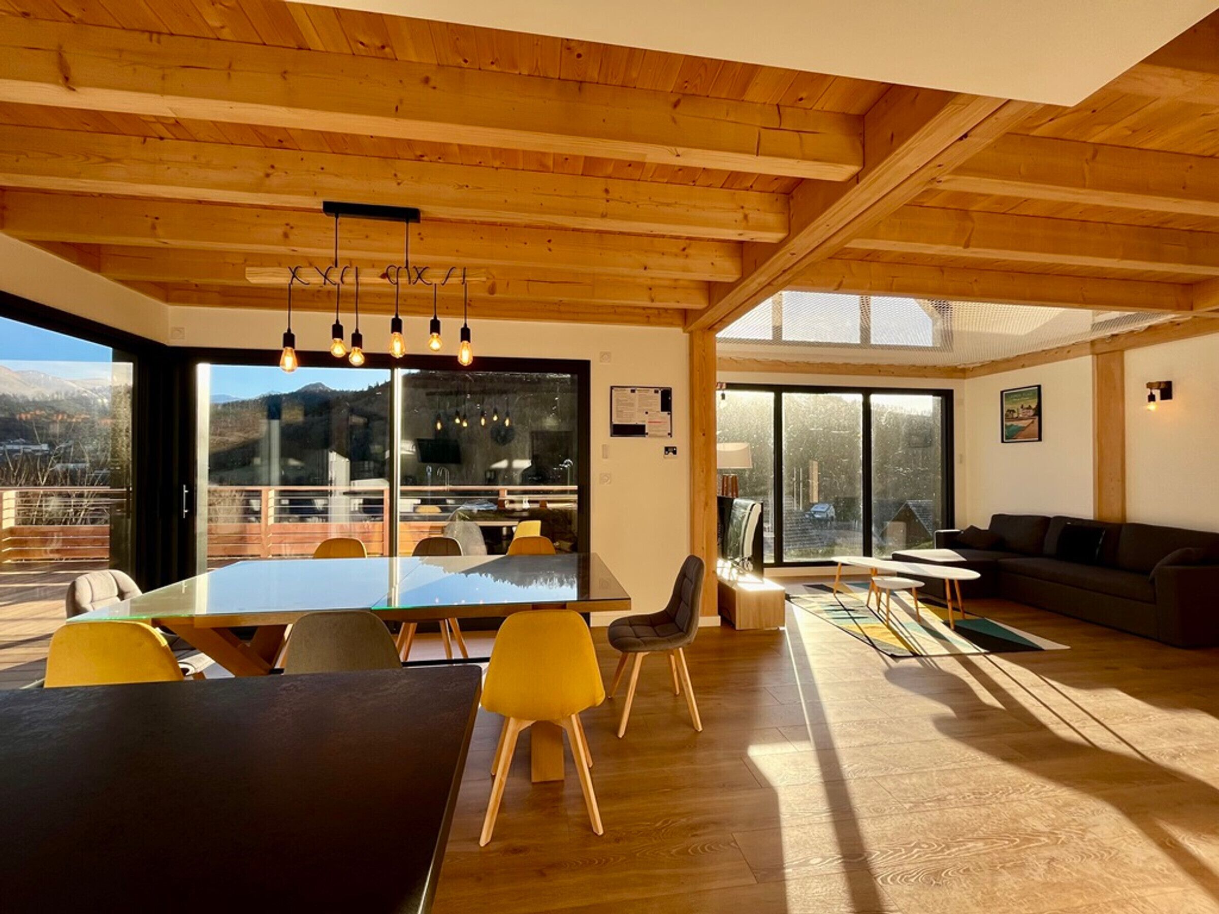A spacious dining area with a large table and chairs, a wooden ceiling, and large windows offering a mountain view.