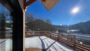 Een houten terras met een met sneeuw bedekte grond, een houten leuning en een helder uitzicht op besneeuwde bergtoppen onder een felle zon.