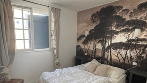 Un dormitorio con cama, una silla, un ventanal con cortinas y un mural en la pared.
