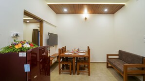 A compact dining area with a wooden table and chairs, a sofa, and a refrigerator with a floral arrangement. - Grand Plaza Inn (Tirur)