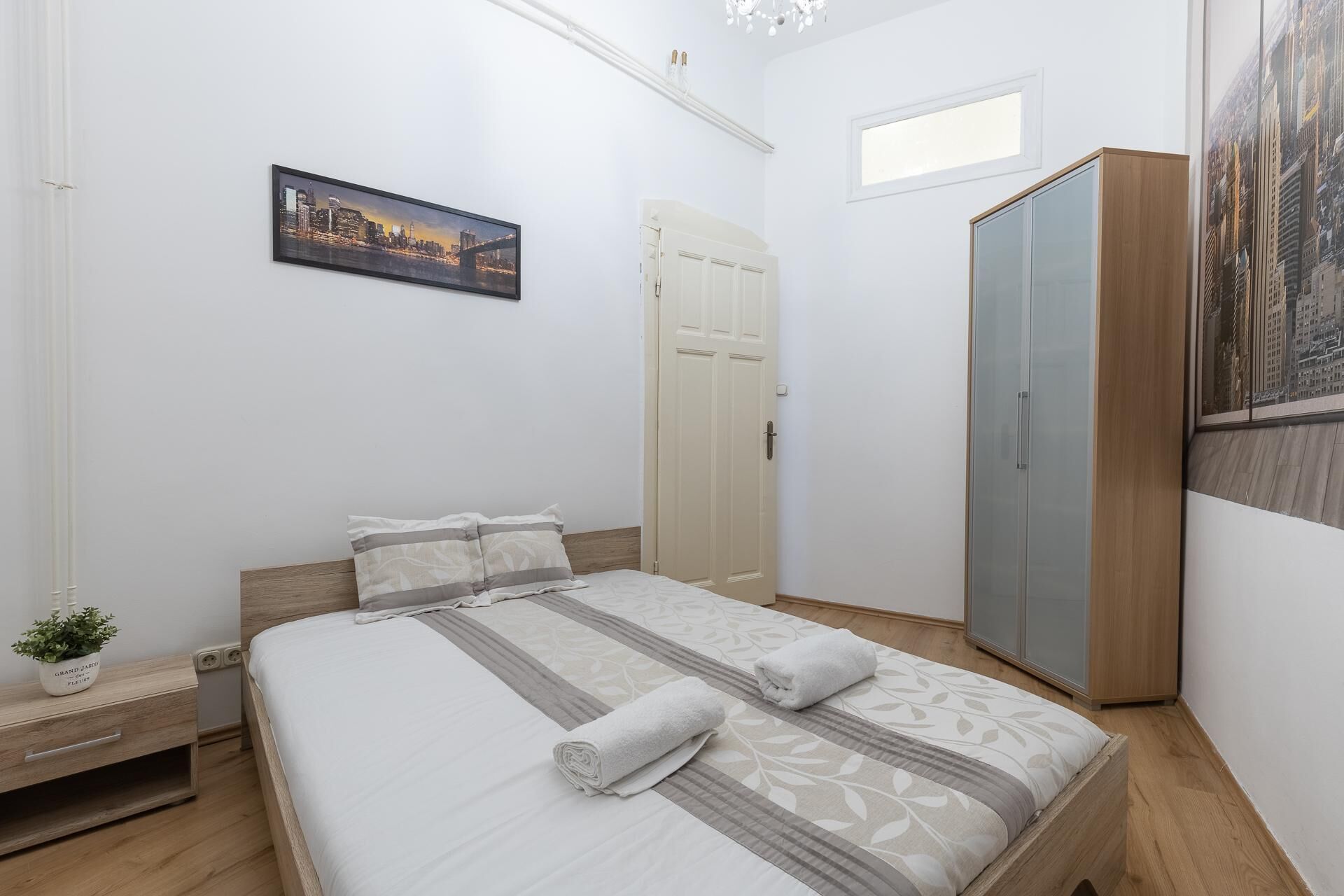 Un dormitorio ordenado con cama, armario, mesita de noche y un cuadro de un paisaje urbano en la pared.
