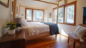 A bedroom with a wooden bed, large windows, a bedside table with a lamp, and a potted plant. - Peaceful and spacious home tucked away at the base of the Mission Mountains. (St. Ignatius)