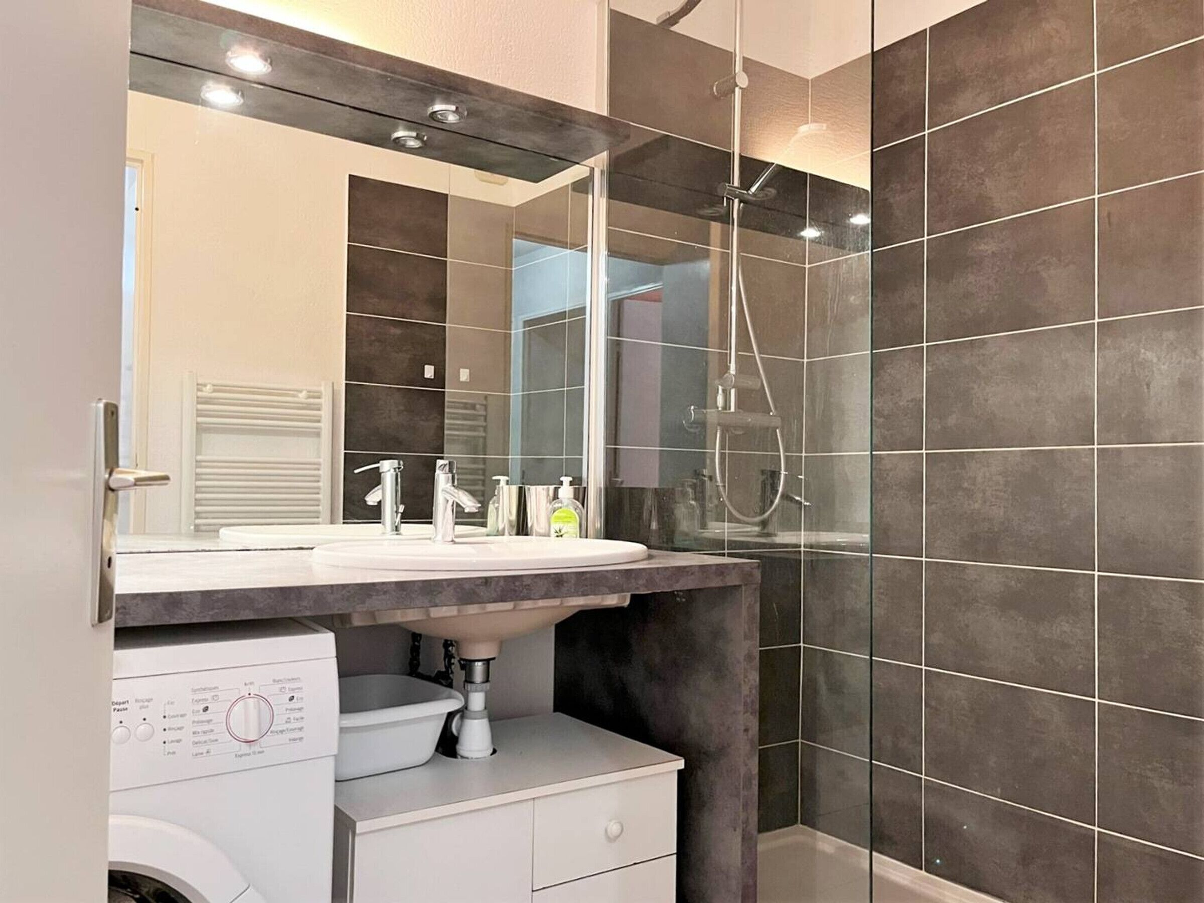 Un baño moderno con un espejo grande, un lavamanos, una ducha y una lavadora.