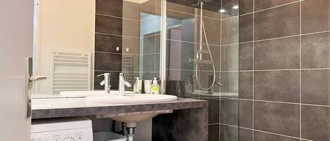 Un baño moderno con un espejo grande, un lavamanos, una ducha y una lavadora.