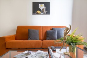 Un salon avec un canapé orange, une table basse en verre, un mannequin en bois et une plante de fougère.