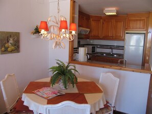 Un espace repas comprenant une table, des chaises, un lustre, une plante en pot et une cuisine visible à travers une porte ouverte.