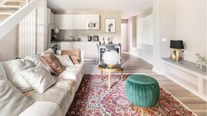 Un salon moderne avec un canapé, un pouf et une table basse. On aperçoit une cuisine en arrière-plan, avec des meubles blancs et un four à micro-ondes.