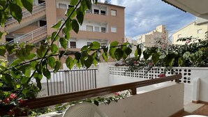 A balcony with white plastic chairs and a wooden railing, overlooking a residential area with multi-story buildings and a white fence.