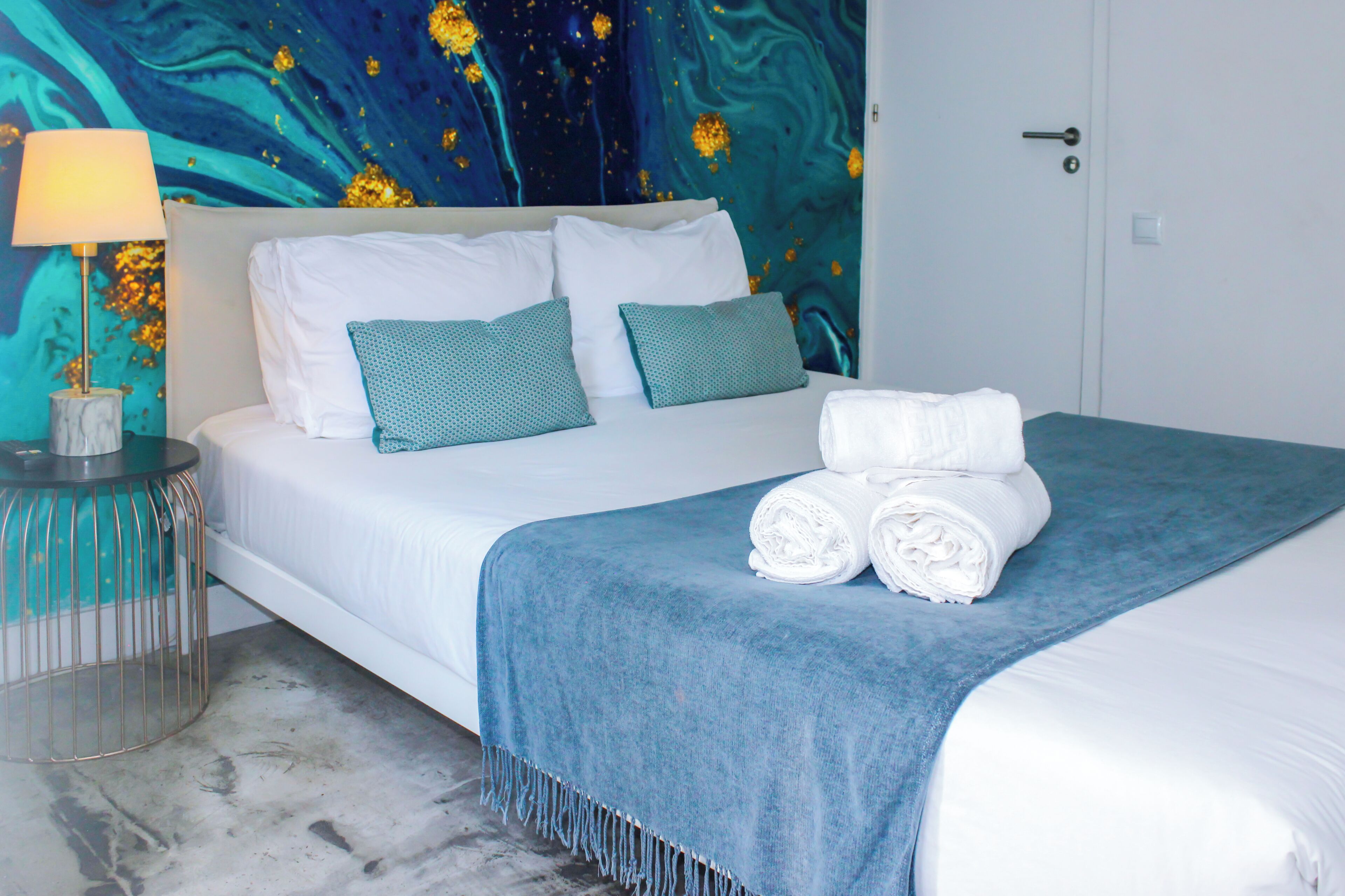 A neatly made bed with white linens and teal pillows, a blue throw blanket, and rolled towels on the side.