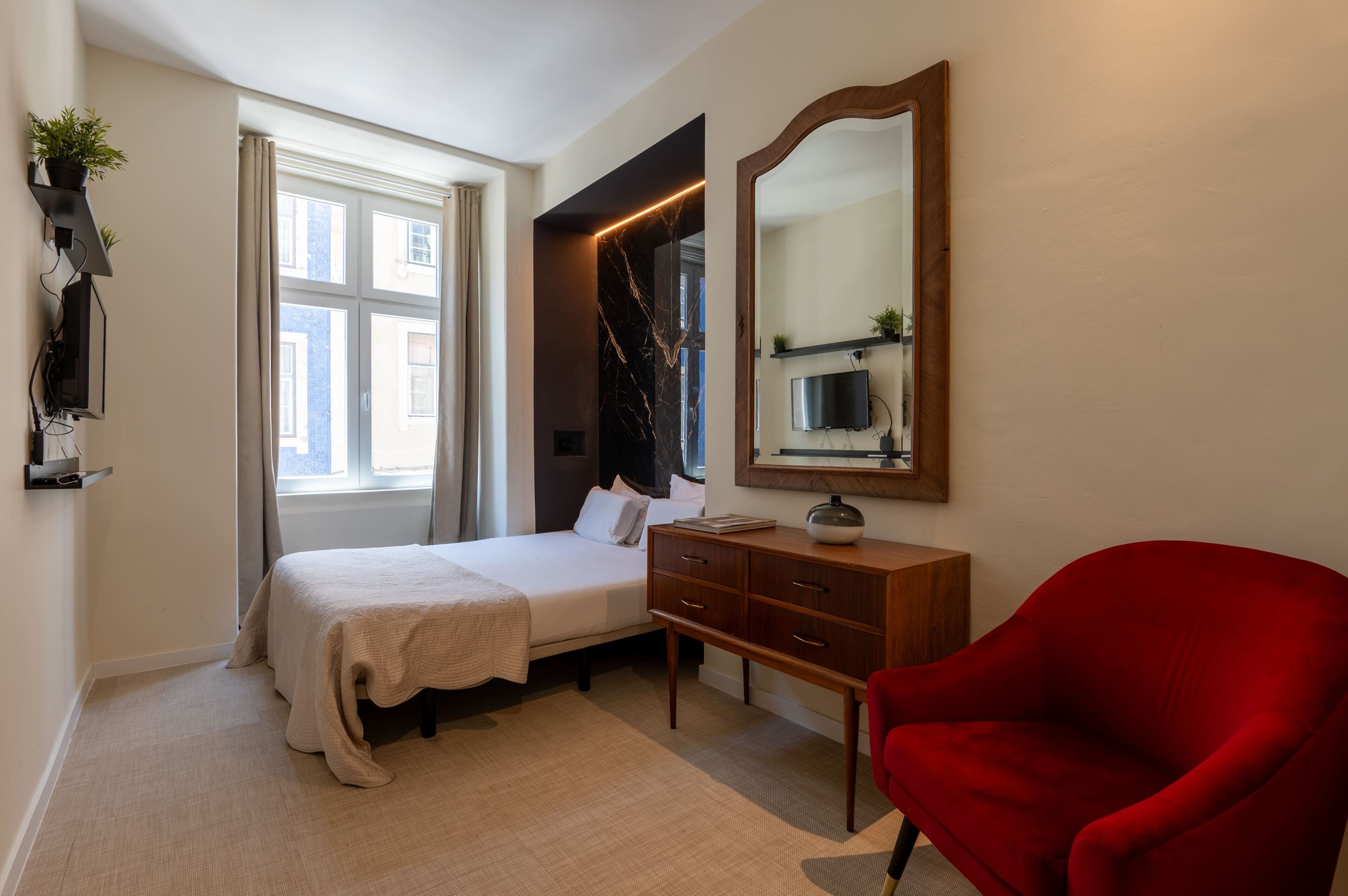 A bedroom with a bed, a wooden dresser, a red chair, a mirror, and a TV on the wall.