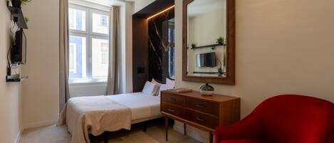 A bedroom with a bed, a wooden dresser, a red chair, a mirror, and a TV on the wall.