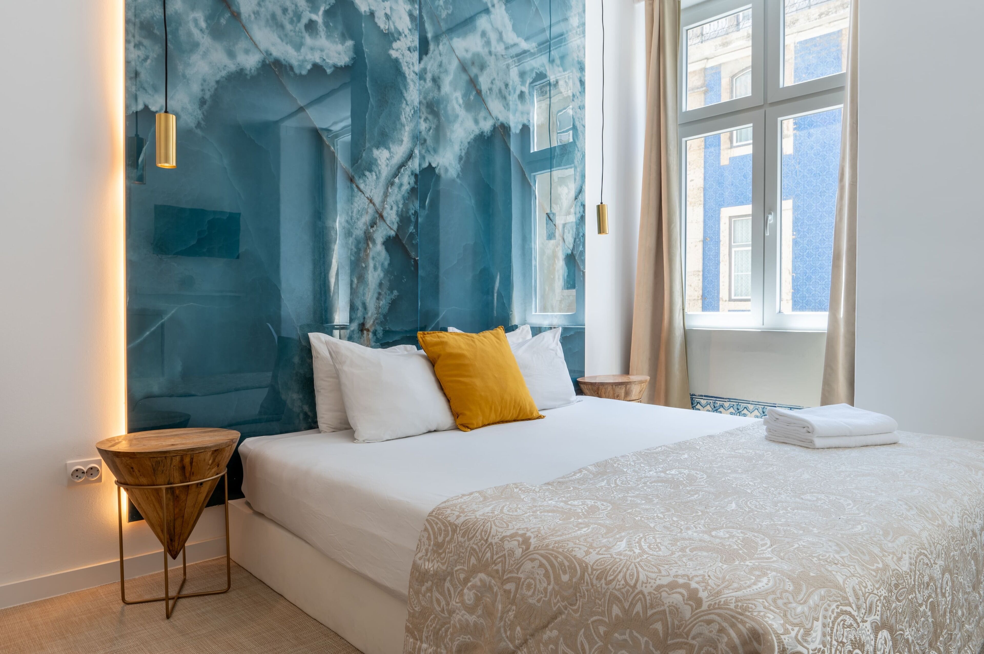 A bedroom with a bed, a bedside table, a wall with a blue abstract design, and a window with curtains.