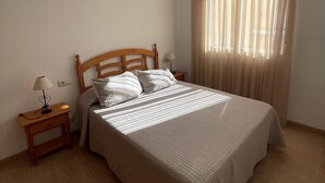 A bedroom with a wooden headboard, a bed with white bedding, two bedside tables with lamps, and a window with sheer curtains. - Cozy 2-Bed Apartment, 150m from Beach, Pool, Parking, Peníscola (Peñíscola)