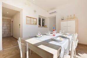 Un coin repas avec une longue table dressée pour quatre personnes, des chaises blanches, une fenêtre donnant sur un beau paysage et un buffet rempli de bouteilles.