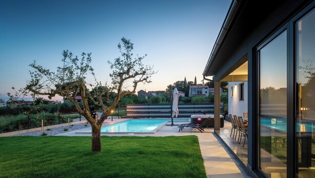 Una casa moderna con piscina, césped cuidado y zona de patio con mobiliario exterior.