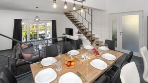 Un comedor moderno con una mesa de madera preparada para seis comensales, una escalera y un sofá al fondo.