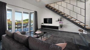 Una sala de estar moderna con un sofá gris oscuro, una mesa de centro de vidrio y un televisor negro en la pared. Una escalera de madera con barandilla negra conduce a un nivel superior.