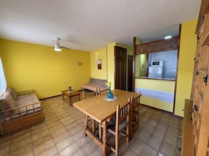 A dining area with a wooden table and chairs, a sofa, a small table with a vase, and a kitchen visible through a partition.