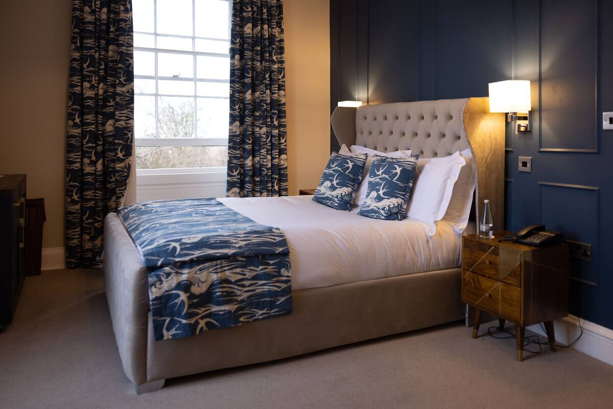 A bedroom with a tufted headboard, a bedside lamp, a telephone, and a window with curtains.