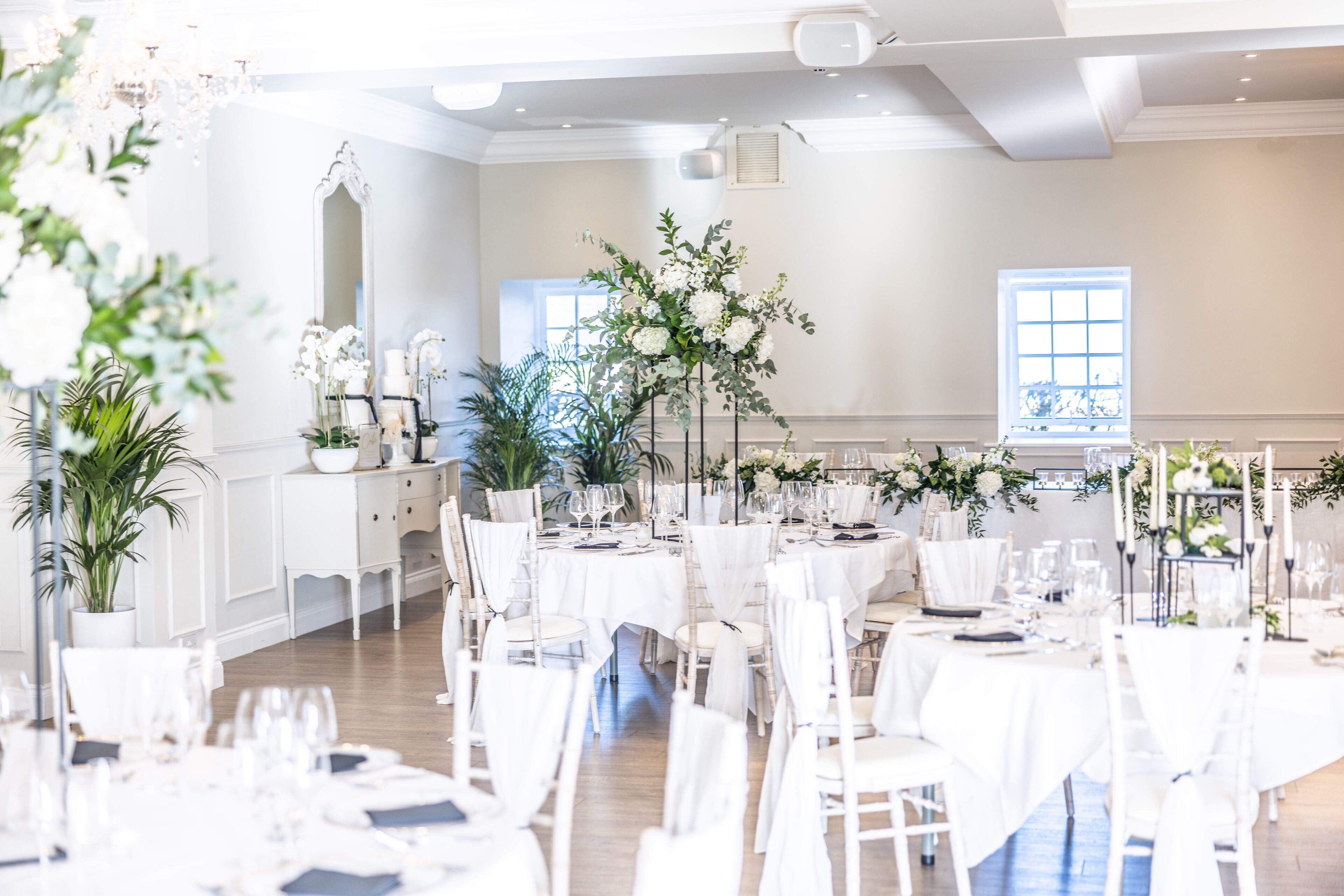 A spacious room with multiple tables set for dining, adorned with white tablecloths and floral centerpieces.