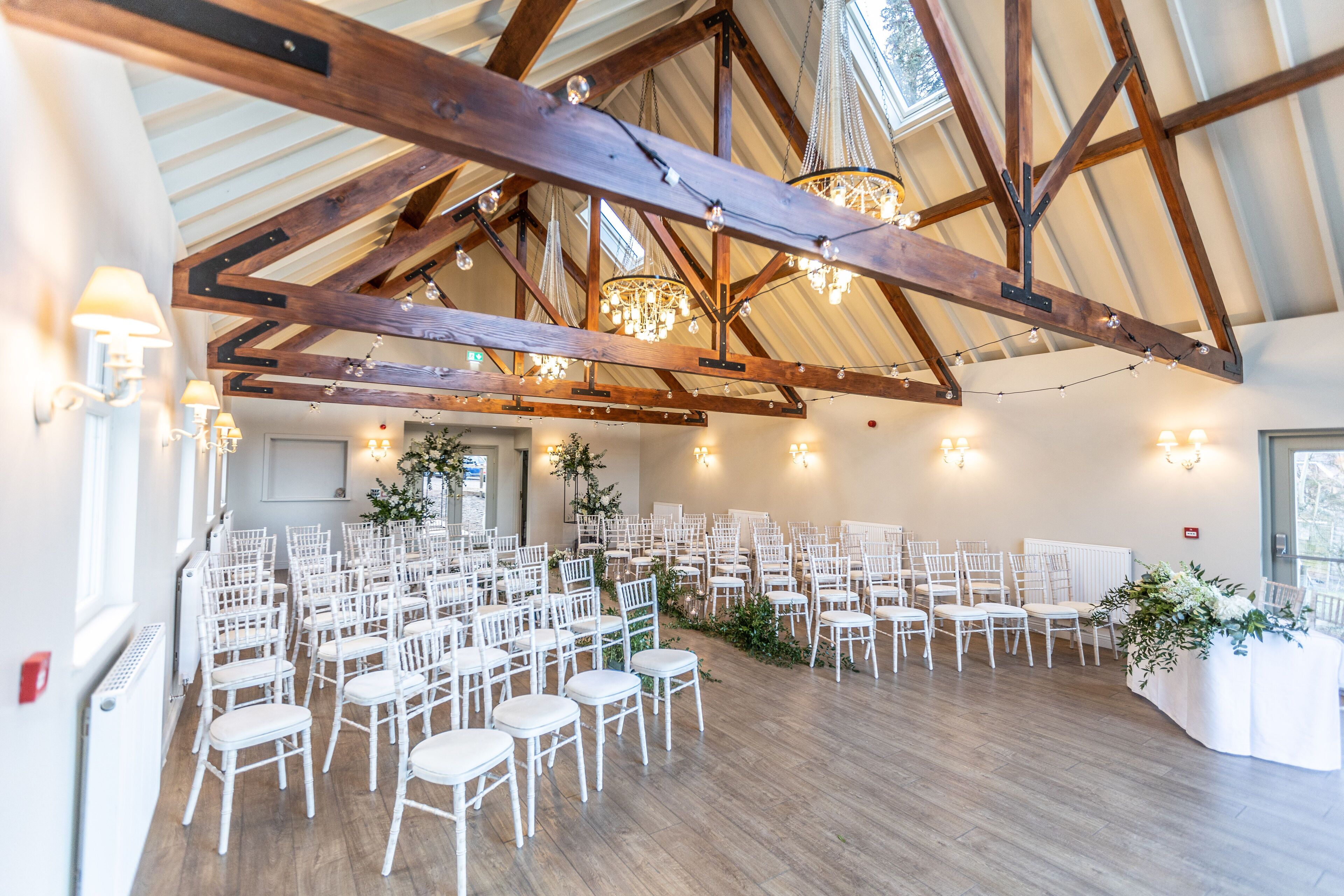 A spacious hall with wooden beams and chandeliers, arranged seating, and a white table with floral arrangements.