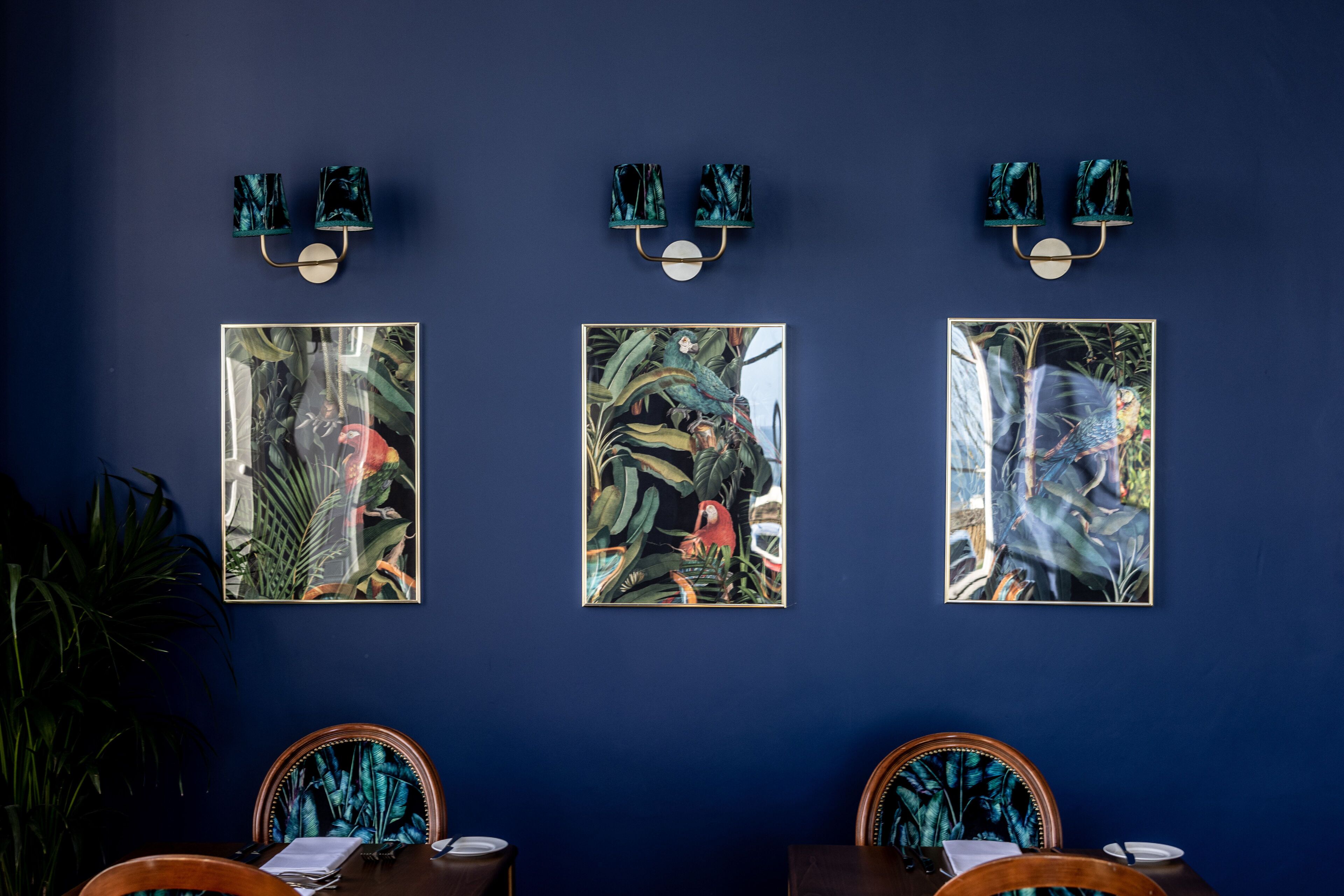 Zona pranzo con una parete blu scuro, tre opere d'arte tropicali incorniciate e due sedie di legno con cuscini coordinati.