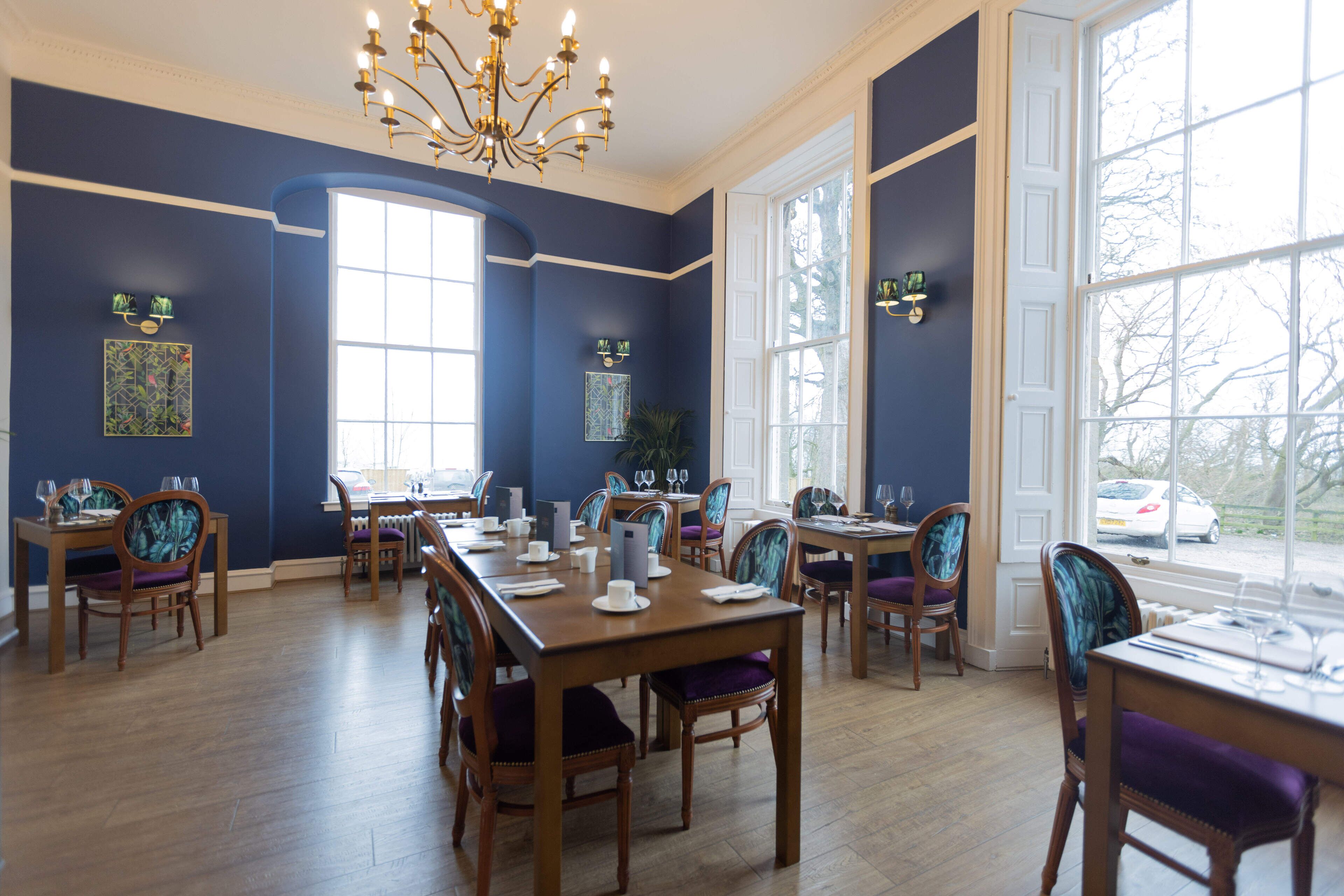 A dining area with multiple tables and chairs, large windows, and a chandelier.