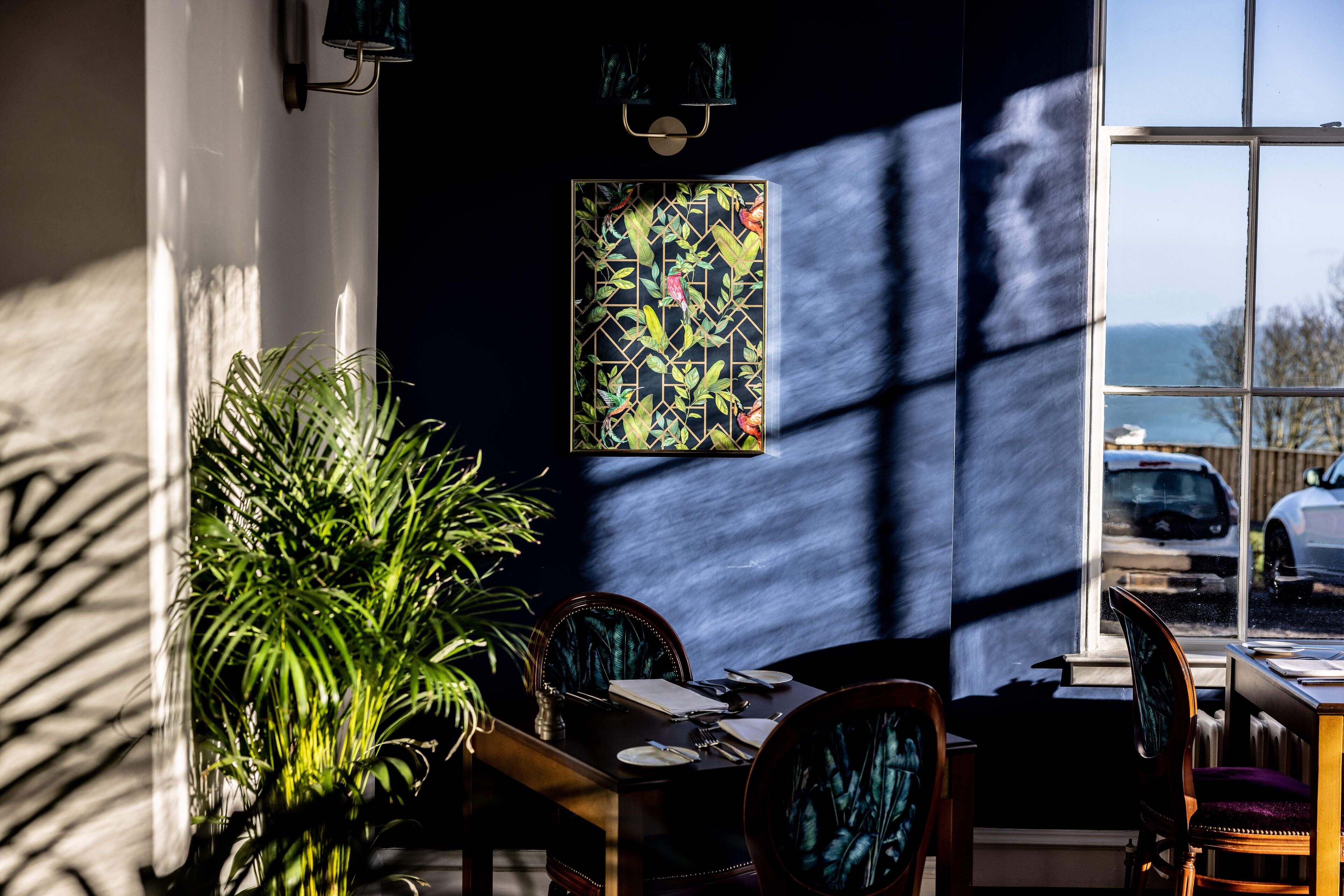 A dining area with a table set for two, a large potted plant, and a view of the sea through a window.