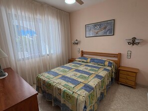 A bedroom with a bed, a nightstand, a lamp, a window with curtains, and a wall-mounted light fixture. - Apartment in Peniscola with pool, tennis court, and playground (Peñíscola)