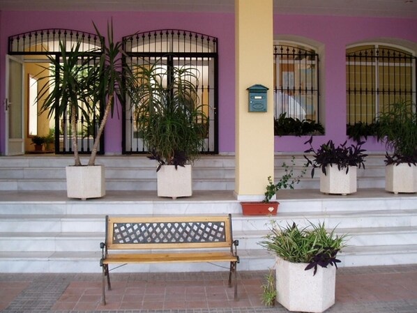 Un banco frente a un edificio de fachada morada, plantas en macetas y un buzón.