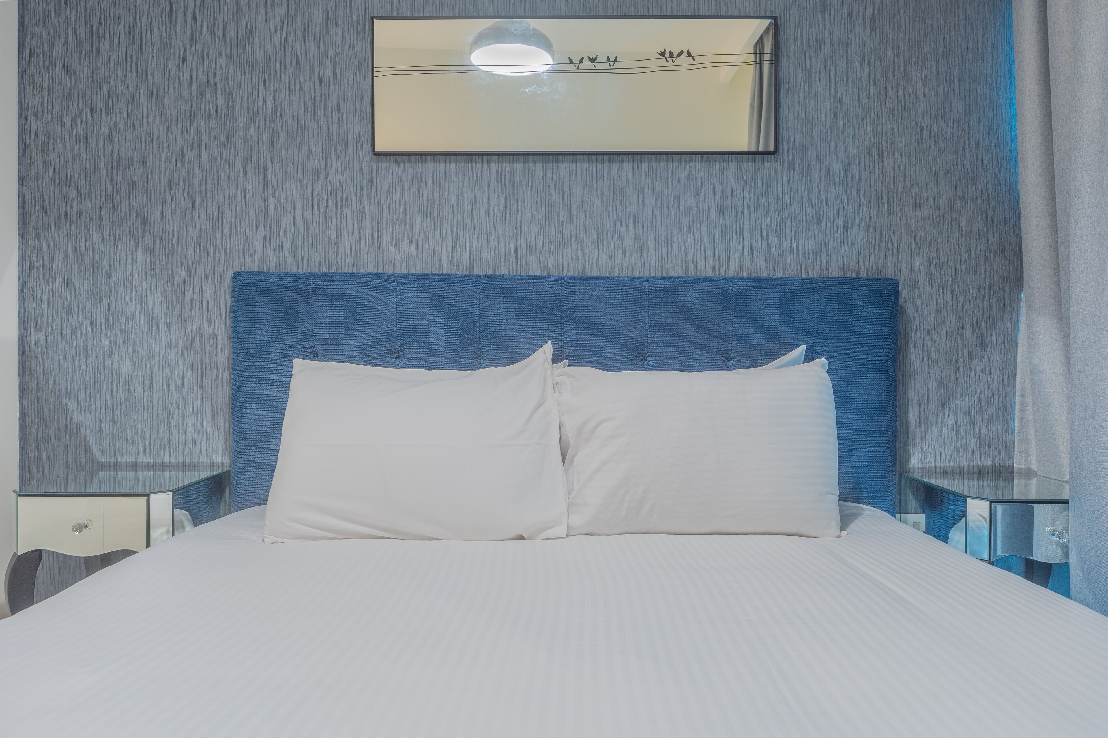Una cama bien hecha con sábanas blancas y dos almohadas, un cabecero azul y una mesita de noche con una lámpara.
