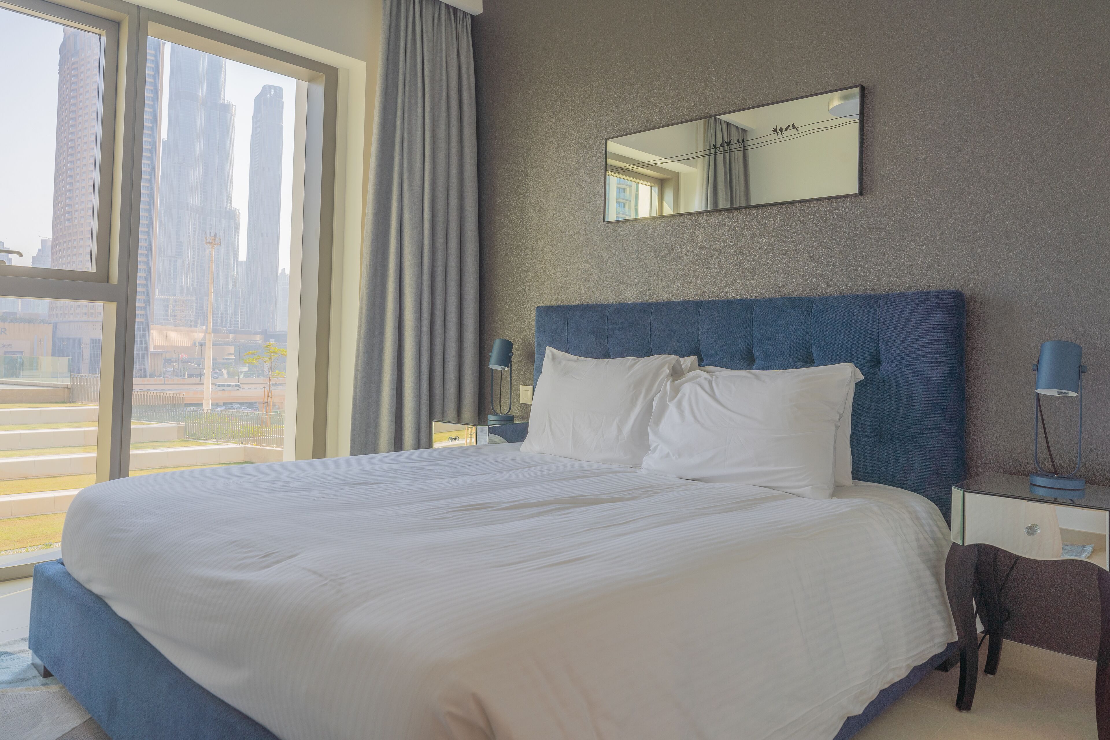 A hotel room with a large bed, a nightstand, a chair, a desk, and a window with a city view.