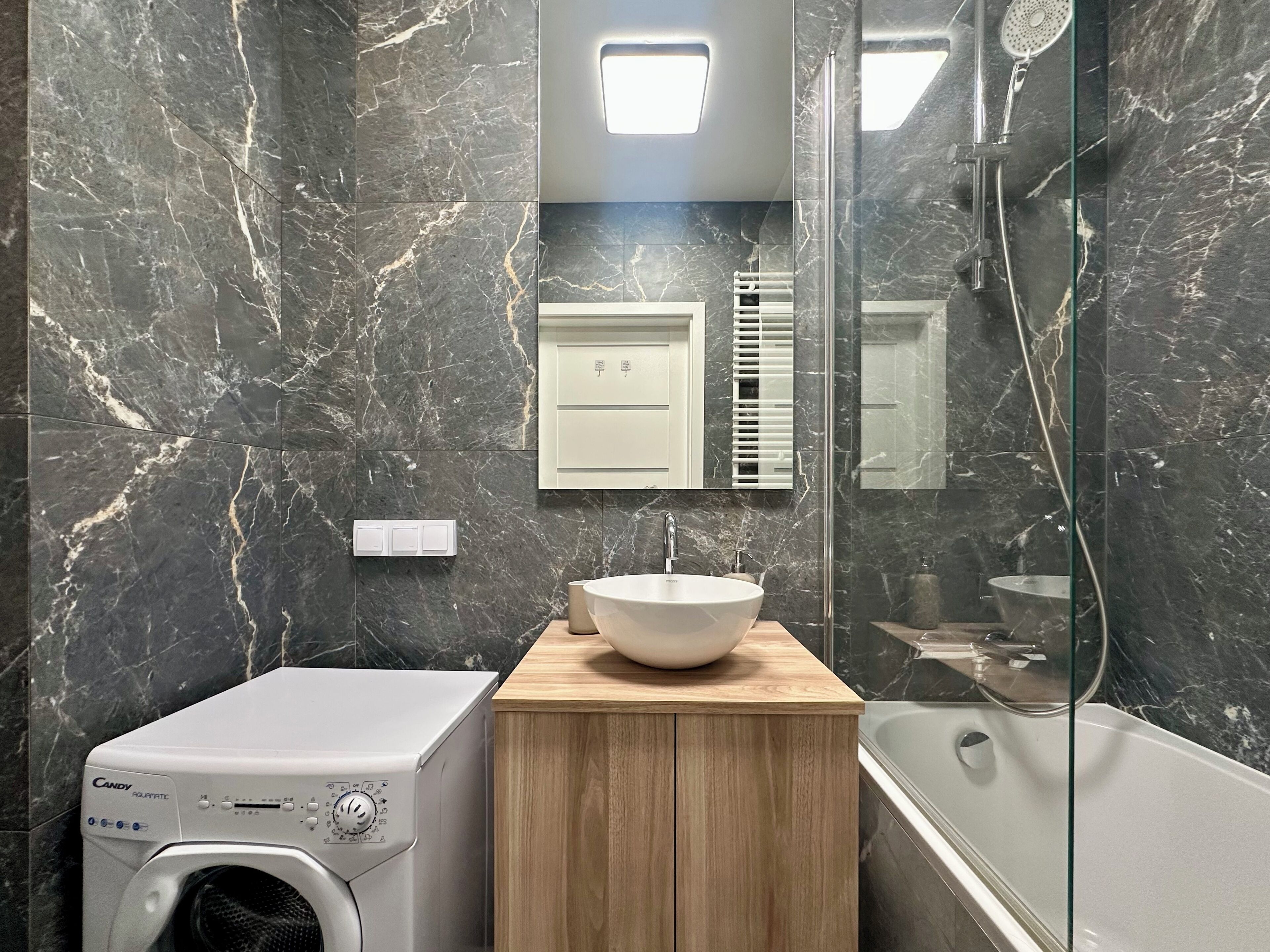 Een moderne badkamer met een marmeren wand, een houten wastafelmeubel met een wit keramieken wastafel, een ingebouwde wasmachine en een douchecabine met glazen wanden.
