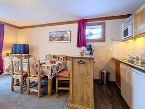 Uma área de refeições acolhedora, com uma mesa e cadeiras de madeira, uma cozinha compacta com micro-ondas e uma janela com cortinas.