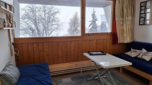 Une pièce chaleureuse avec un mur lambrissé en bois, un canapé bleu, une table blanche et une vue sur les arbres à travers de grandes fenêtres.