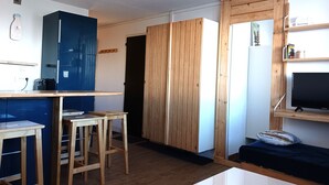 Un espace de vie compact comprenant un coin cuisine, un espace repas avec des tabourets, un téléviseur posé sur un meuble en bois et une porte coulissante donnant sur une autre pièce.