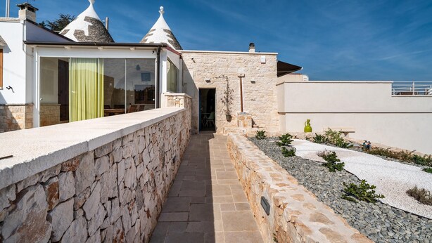 A stone-walled pathway leading to a building with a stone facade and a small garden area with plants and a bench.