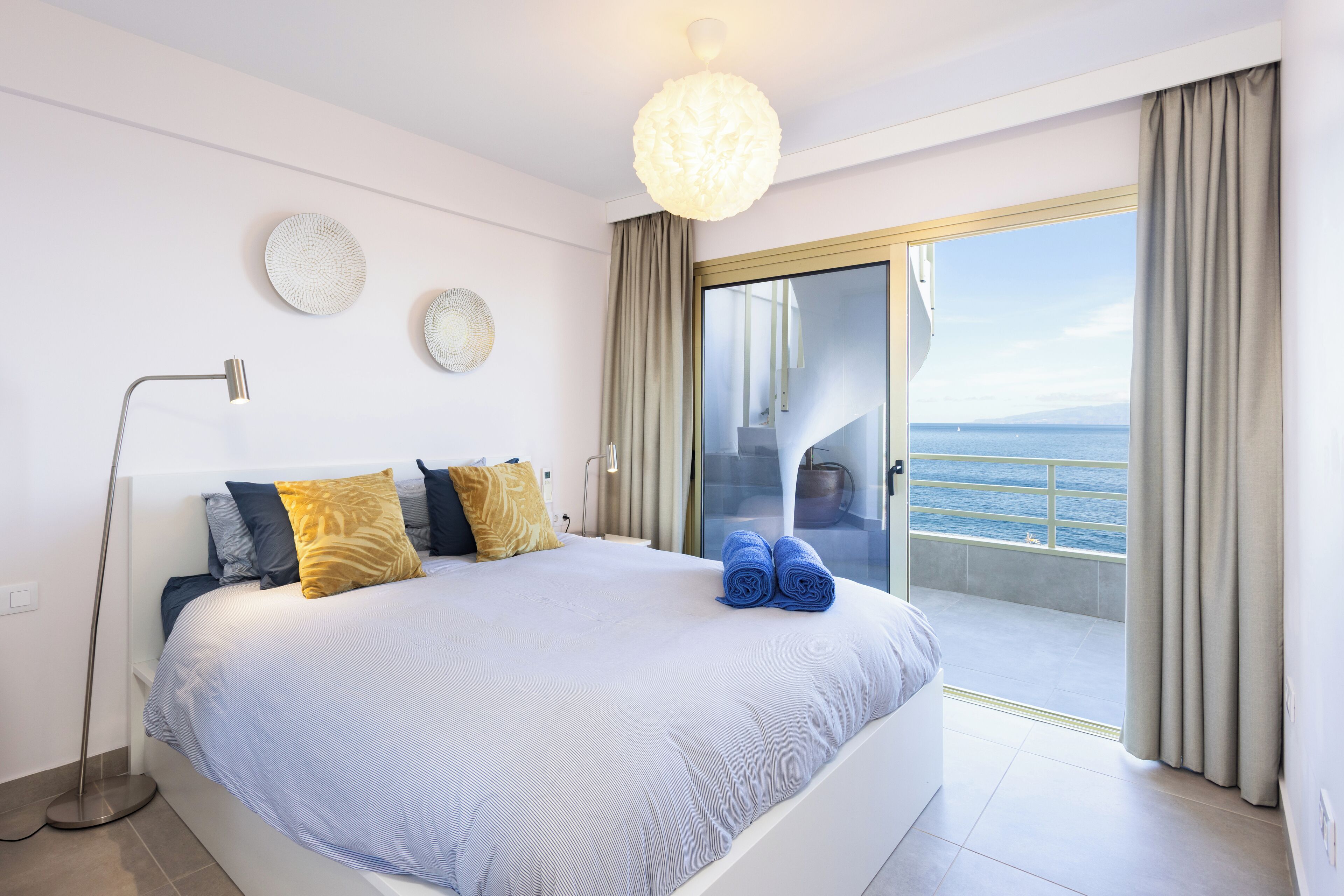 A modern bedroom with a large bed, a floor lamp, and a view of the sea through a sliding glass door.