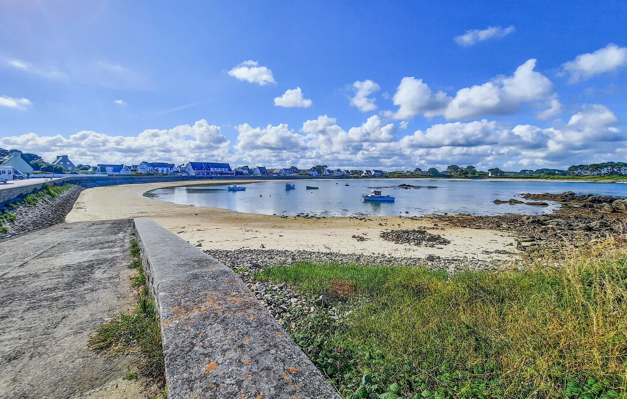 A coastal beach with a curved stone wall, calm water, and boats.