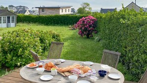 A round wooden table set outdoors with plates, bowls, and food items, surrounded by greenery and a pink flowering bush. - Awesome home in Plouguerneau with house sea view (Plouguerneau)